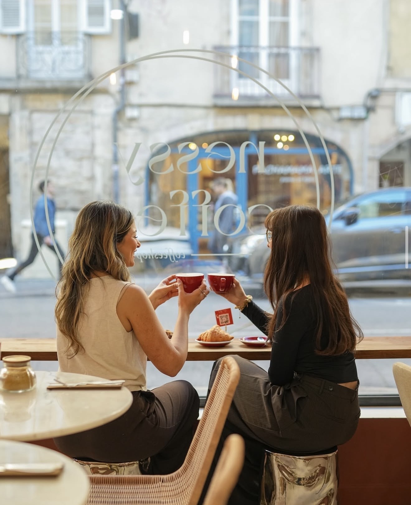 Deux clientes trinquant avec leurs tasses rouges devant la vitrine Nessy Coffee